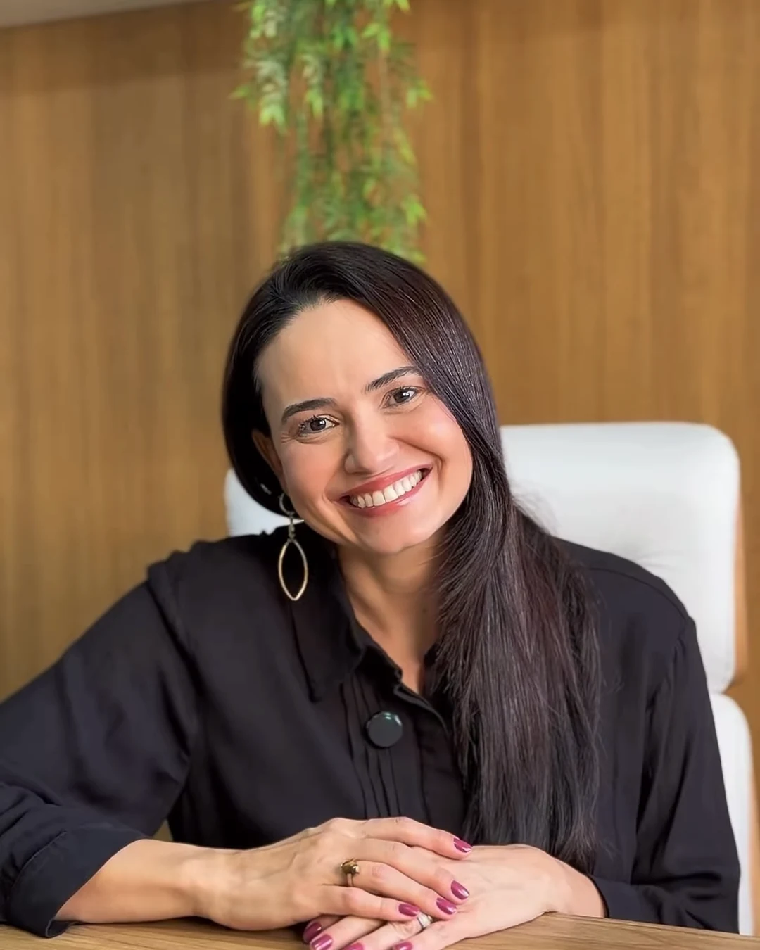 Dra. Luana Menezes sorrindo, apoiada na mesa do consultório, com a parede de madeira e a planta pendente ao fundo